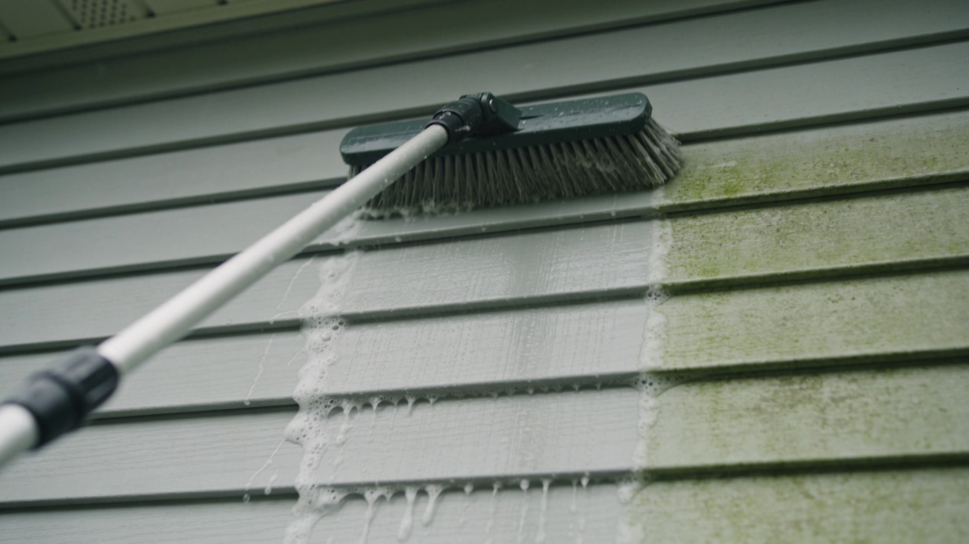 Soft-bristle brush washing pale grey vinyl siding, clean section beside an algae-stained section