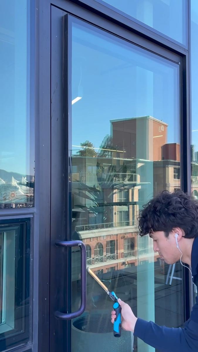 A window cleaner drawing a squeegee down a tall glass door, a brick building reflected in the glass