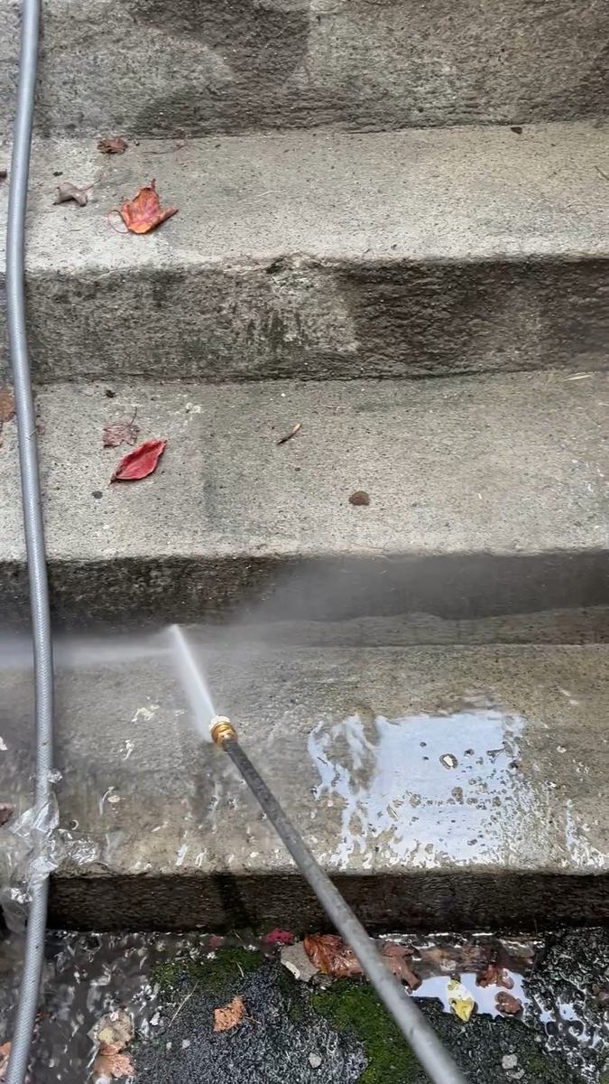 A pressure washer wand spraying a set of grey concrete steps, with the washed strip clearly lighter than the rest